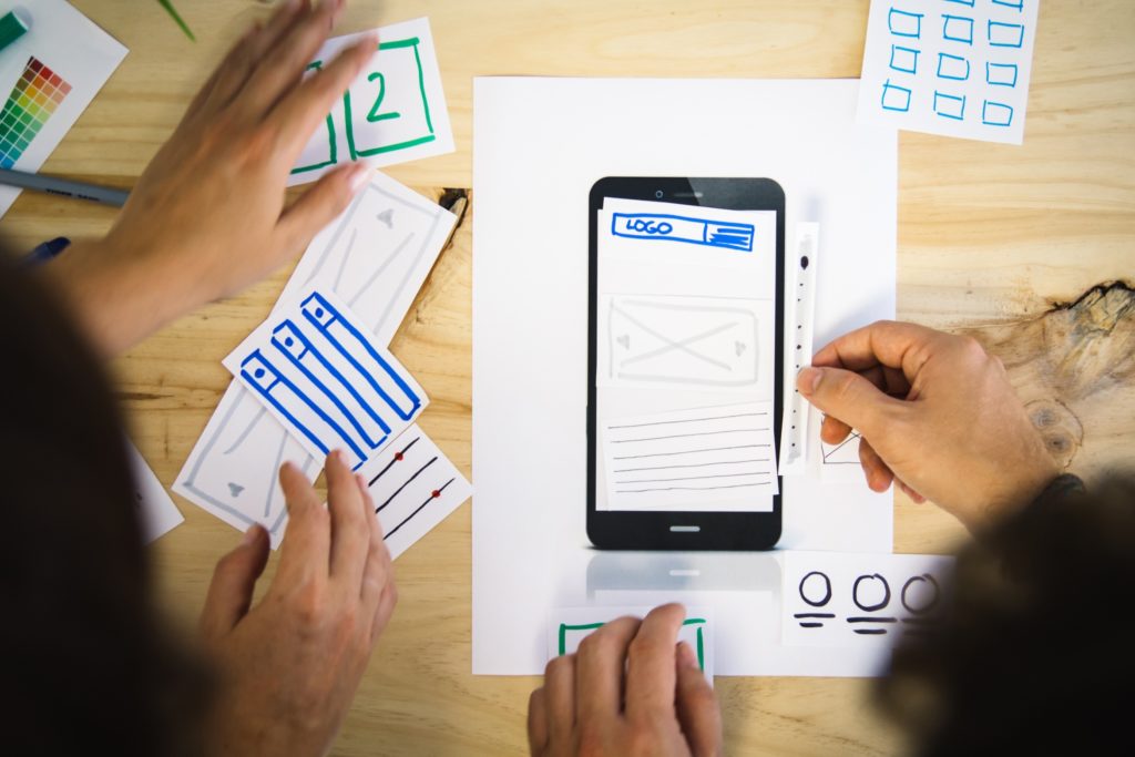 Four hands on table with paper in the shape of a mobile phone user interface to help plan and redesign website experience on mobile. 